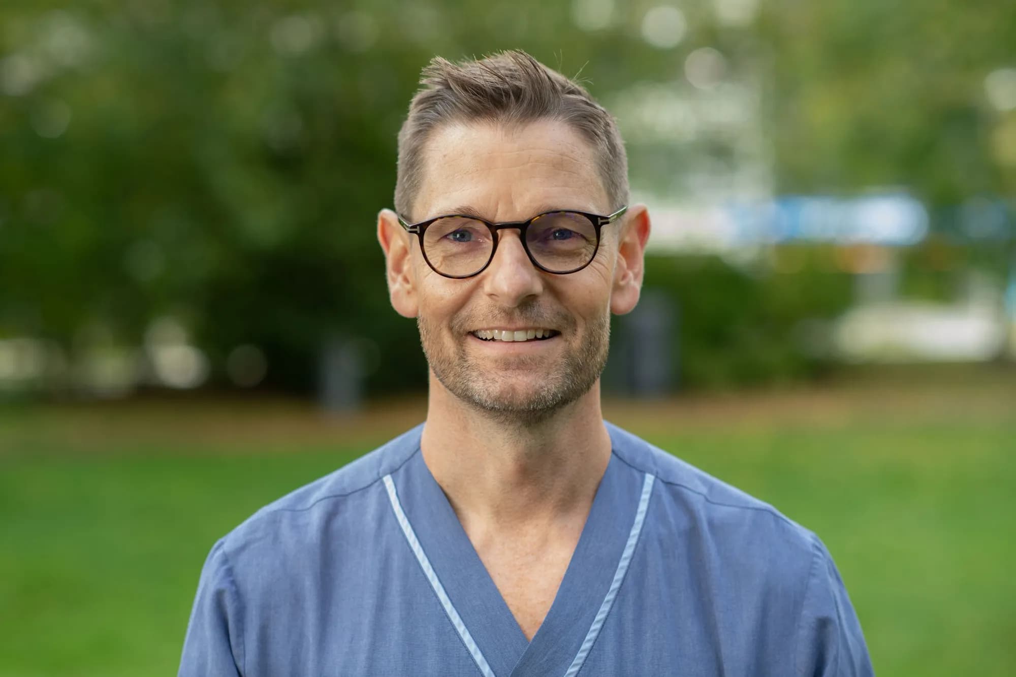 Ulf Kläppe i blå sjukhusskjorta och runda glasögon ler mot kameran utomhus framför grönska.